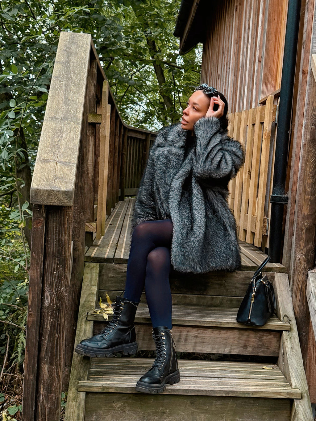 A woman wearing a gray textured coat, navy tights, and black boots sits on the wooden steps of a rustic cabin surrounded by trees and fallen leaves.