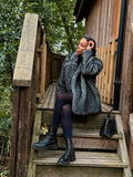 A woman wearing a gray textured coat, navy tights, and black boots sits on the wooden steps of a rustic cabin surrounded by trees and fallen leaves.