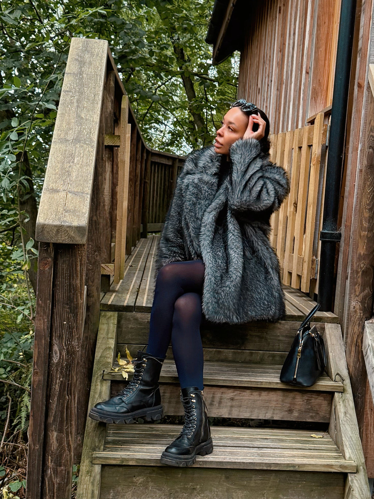 A woman wearing a gray textured coat, navy tights, and black boots sits on the wooden steps of a rustic cabin surrounded by trees and fallen leaves.
