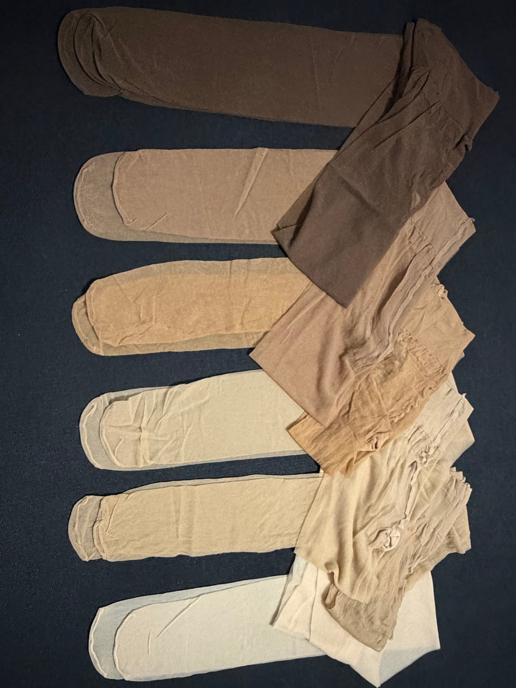 Six pairs of folded tights or pantyhose in different skin-tone shades, arranged vertically on a dark navy background.