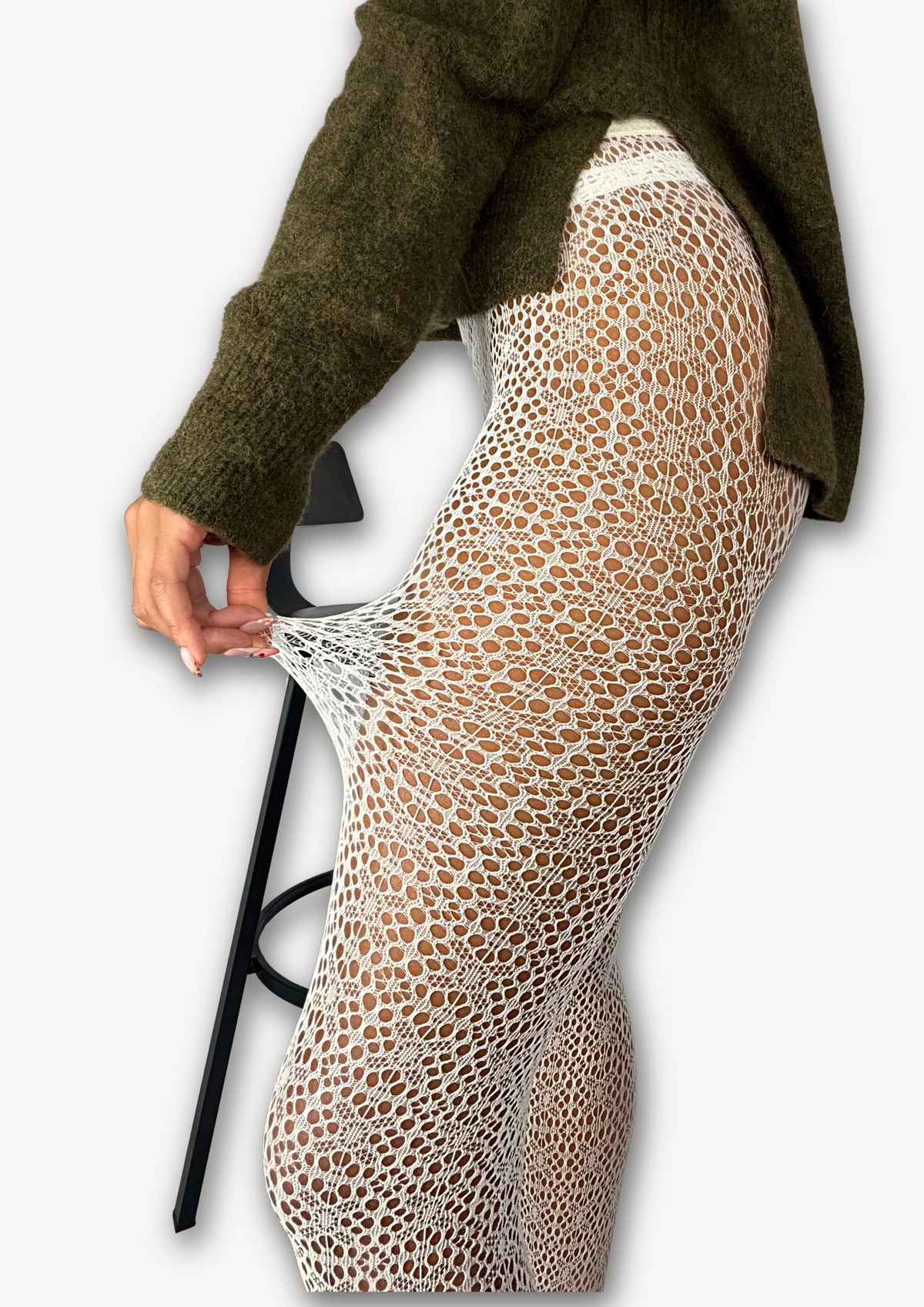 A person wearing a white open-knit, tights with a textured honeycomb pattern, paired with an olive green knit sweater. The person is pulling the skirt slightly outward with one hand to demonstrate its stretch, set against a plain light gray background.